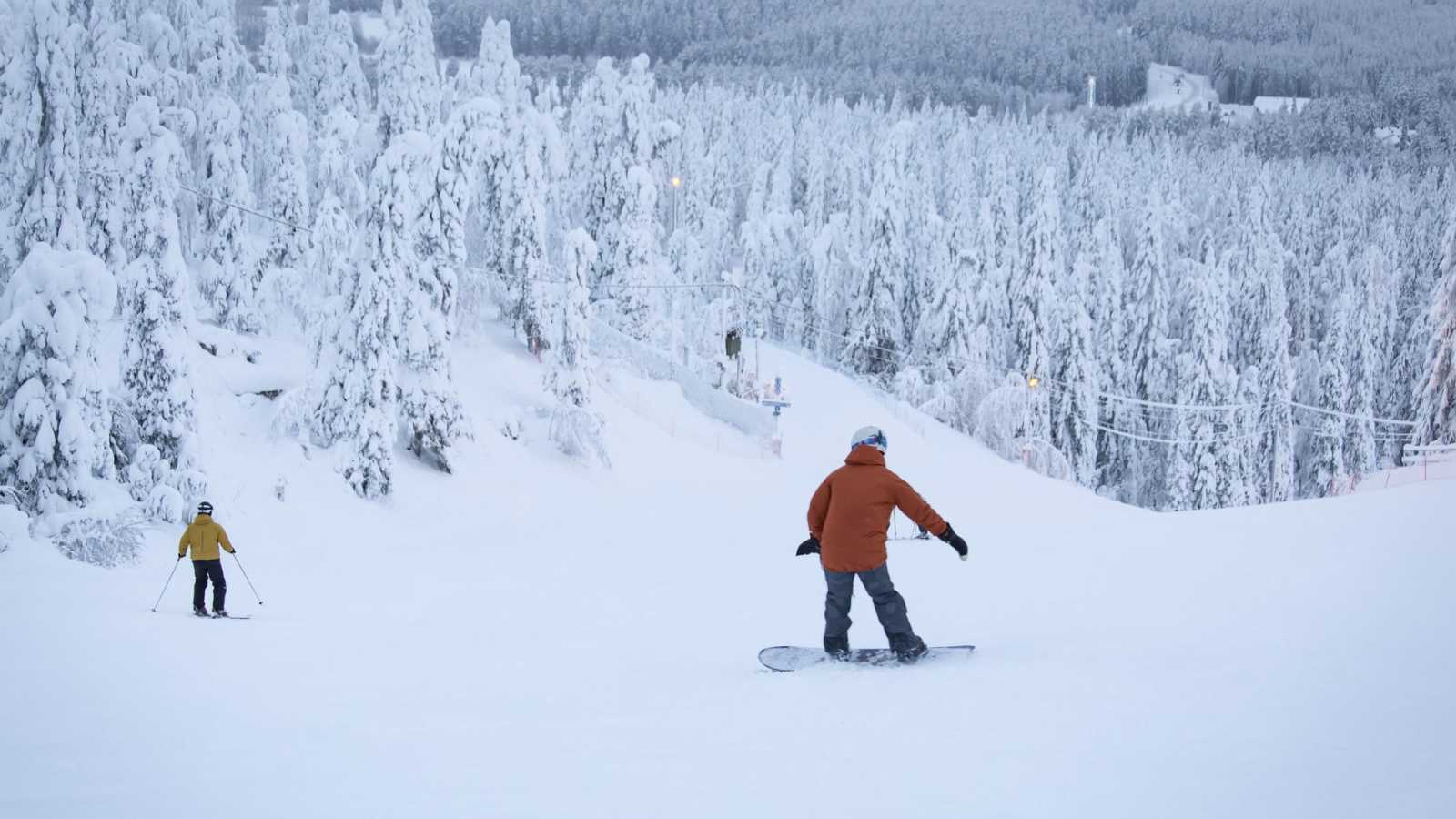Två personer på en snöig skidbacke, en på skidor, den andra på en snowboard.