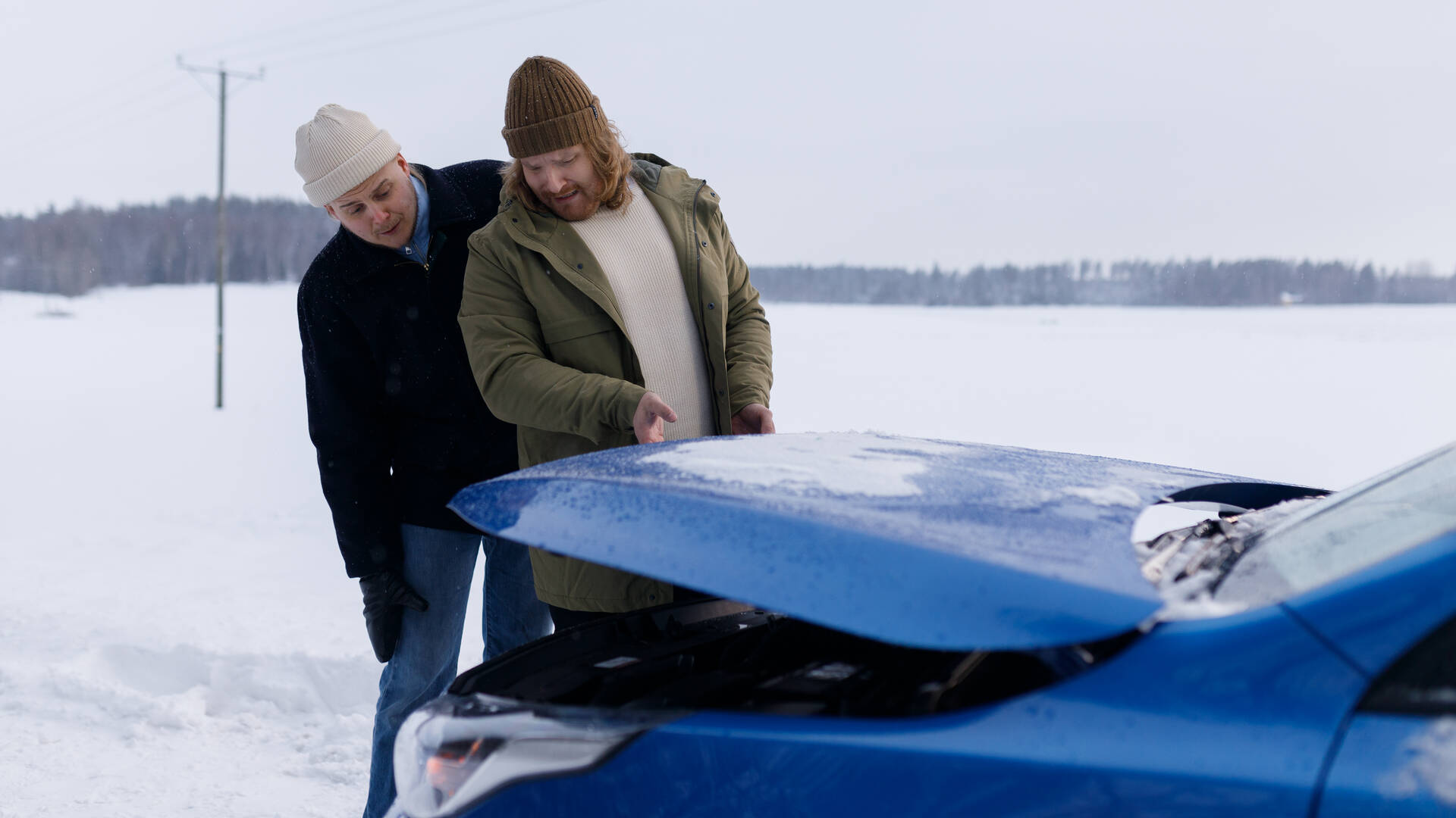 Kaksi miestä on pysähtynyt autolla tielle talvimaisemassa. He nostavat konepeltiä ja katsovat sen alle.