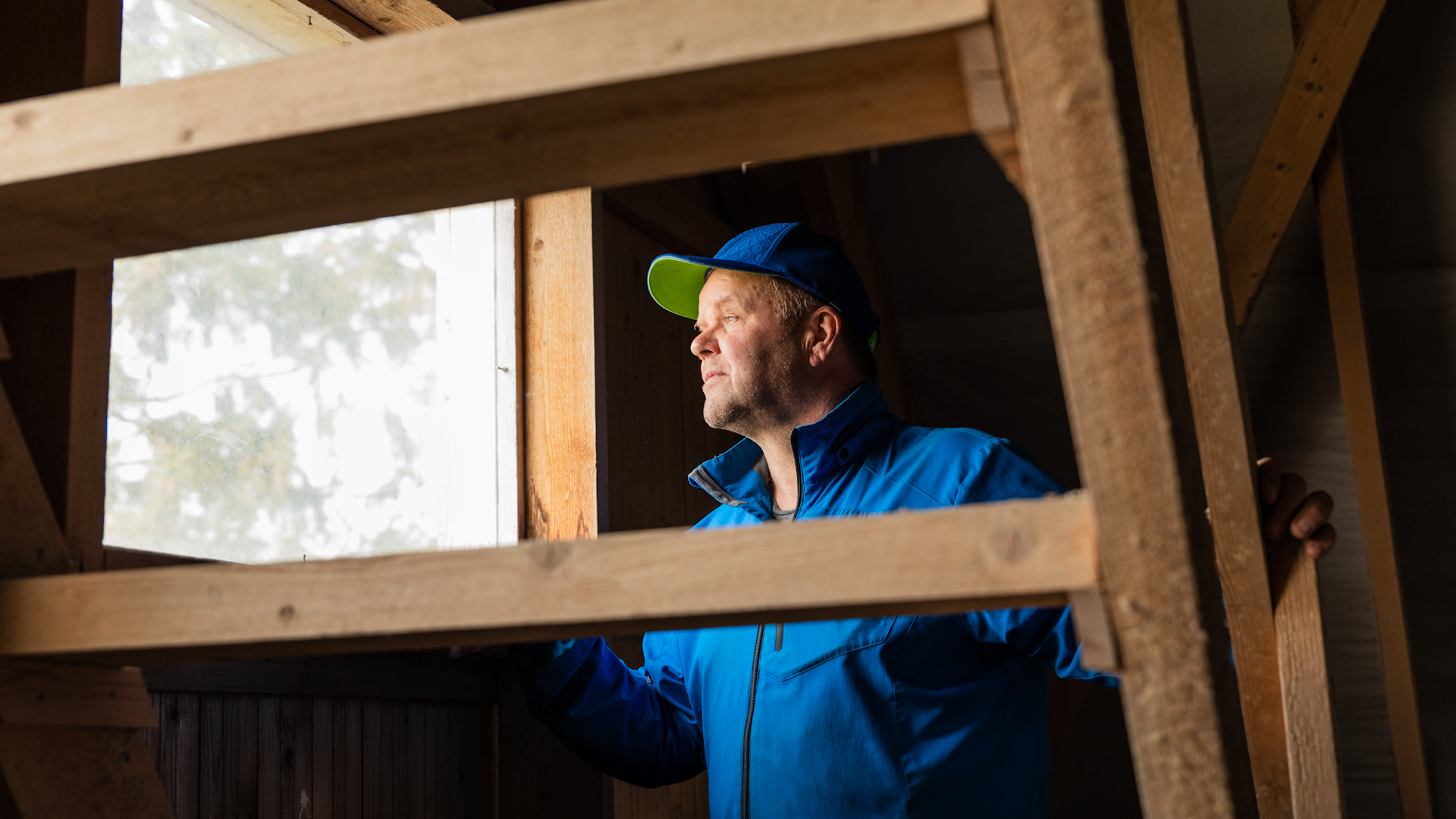 Harmaantunut lippahattupäinen mies katsoo ulos piharakennuksen ikkunasta.