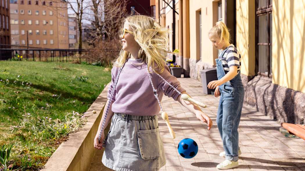 Kaksi nuorta tyttöä leikkii aurinkoisella sisäpihalla. Toisella on hyppynaru, toinen potkii palloa.