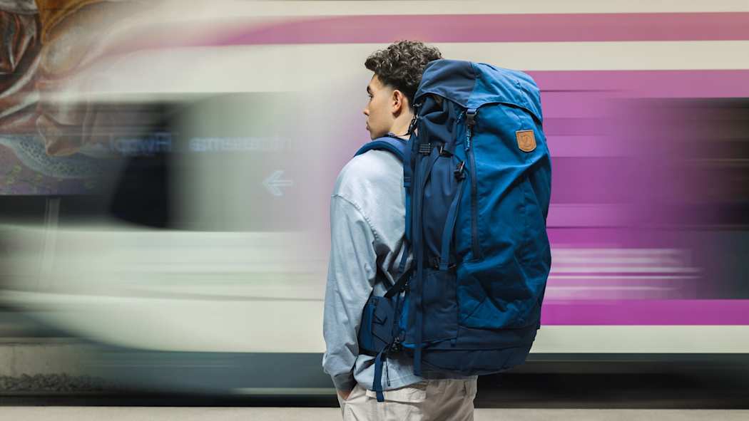 Nuori mies seisoo rinkka selässä, hänen edessään juna kiitää eteenpäin.