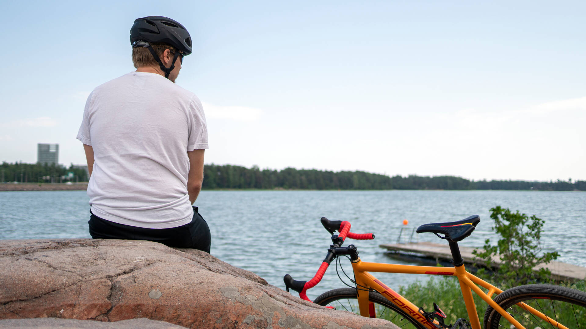Henkilö istuu kalliolla veden äärellä kypärä päässään, vieressä oranssi pyörä nojaa kallioon. Taustalla näkyy puinen laituri ja vastarannalla puita ja rakennuksia.
