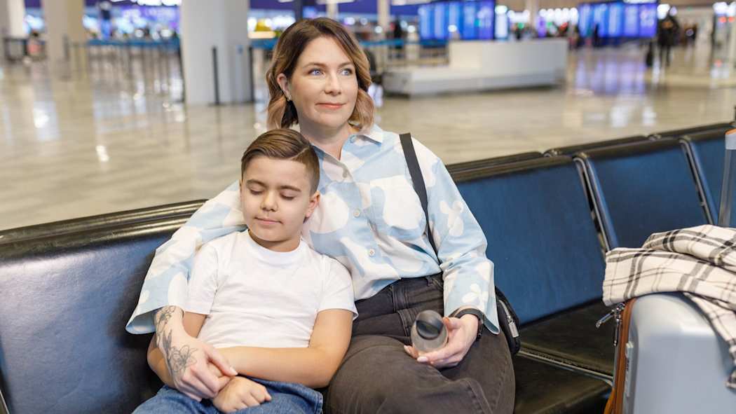 En mamma och hennes son sitter med sitt bagage i väntsalen på flygfältet. En pojke lutar mot sin mamma och sover.