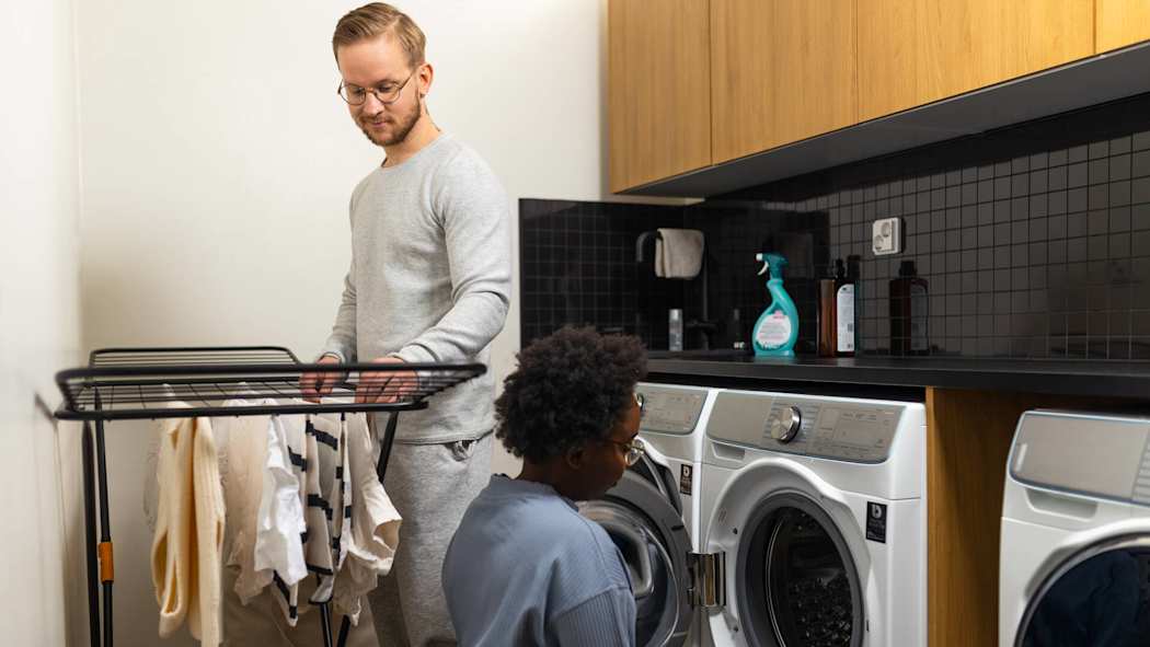 Pariskunta ripustaa pyykkiä kuivumaan. Nainen nostaa pyykkejä koneesta, mies asettelee niitä kuivaustelineelle.