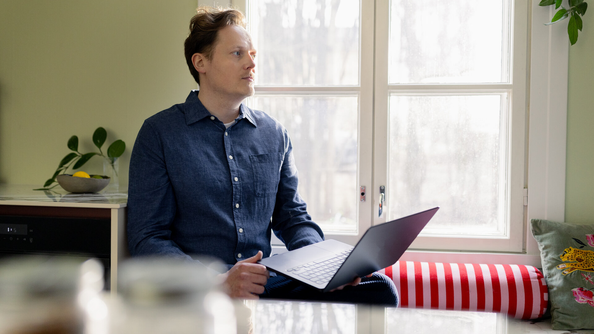 Henkilö istuu kannettava tietokone sylissään ikkunan äärellä. Taustalla näkyy vaaleanvihreä seinä, kasveja ja hedelmäkulho. Ikkunasta tulee runsaasti luonnonvaloa.