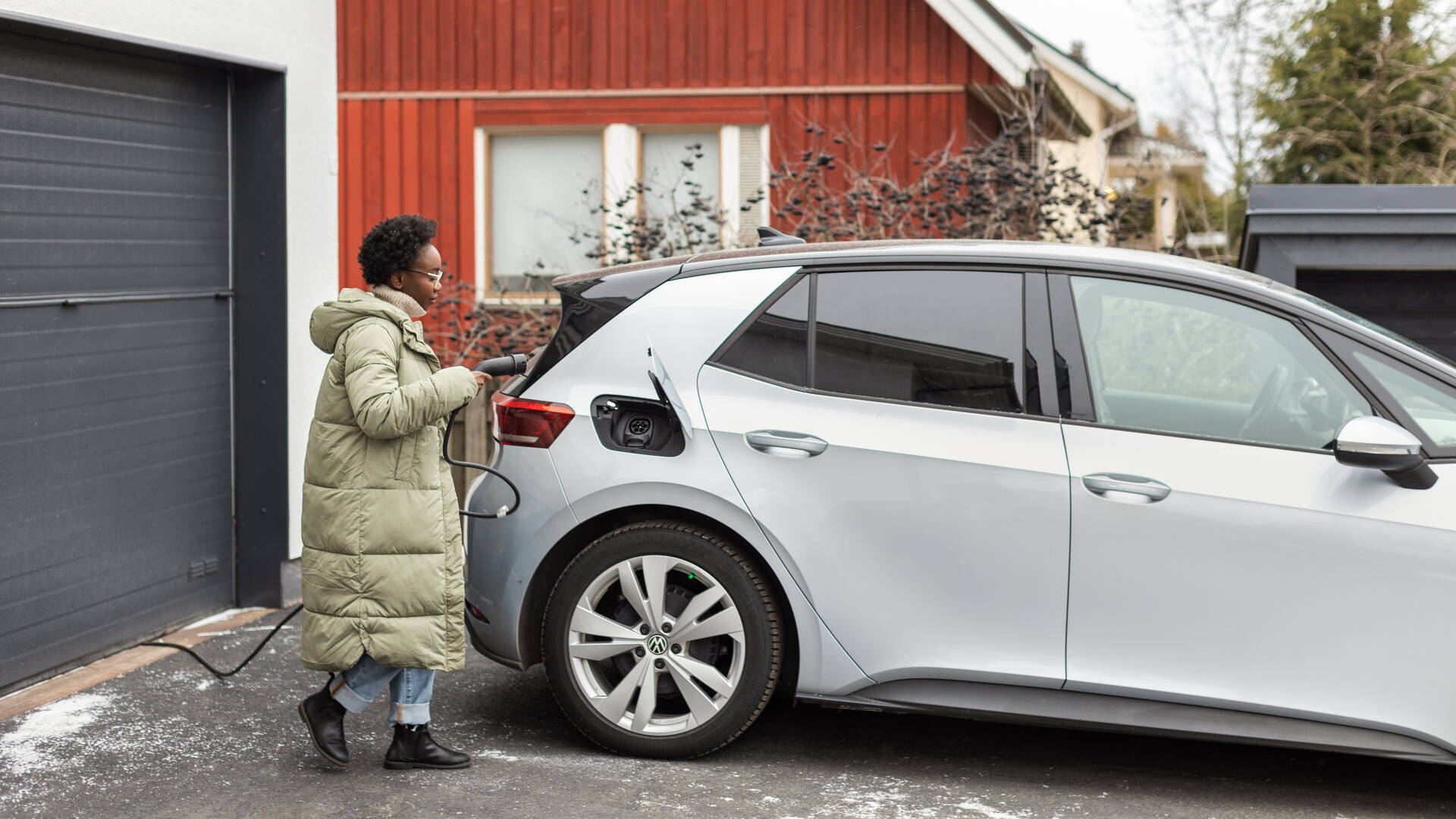 Nainen asettelee laturia sähköautoon autotallinsa edessä. 