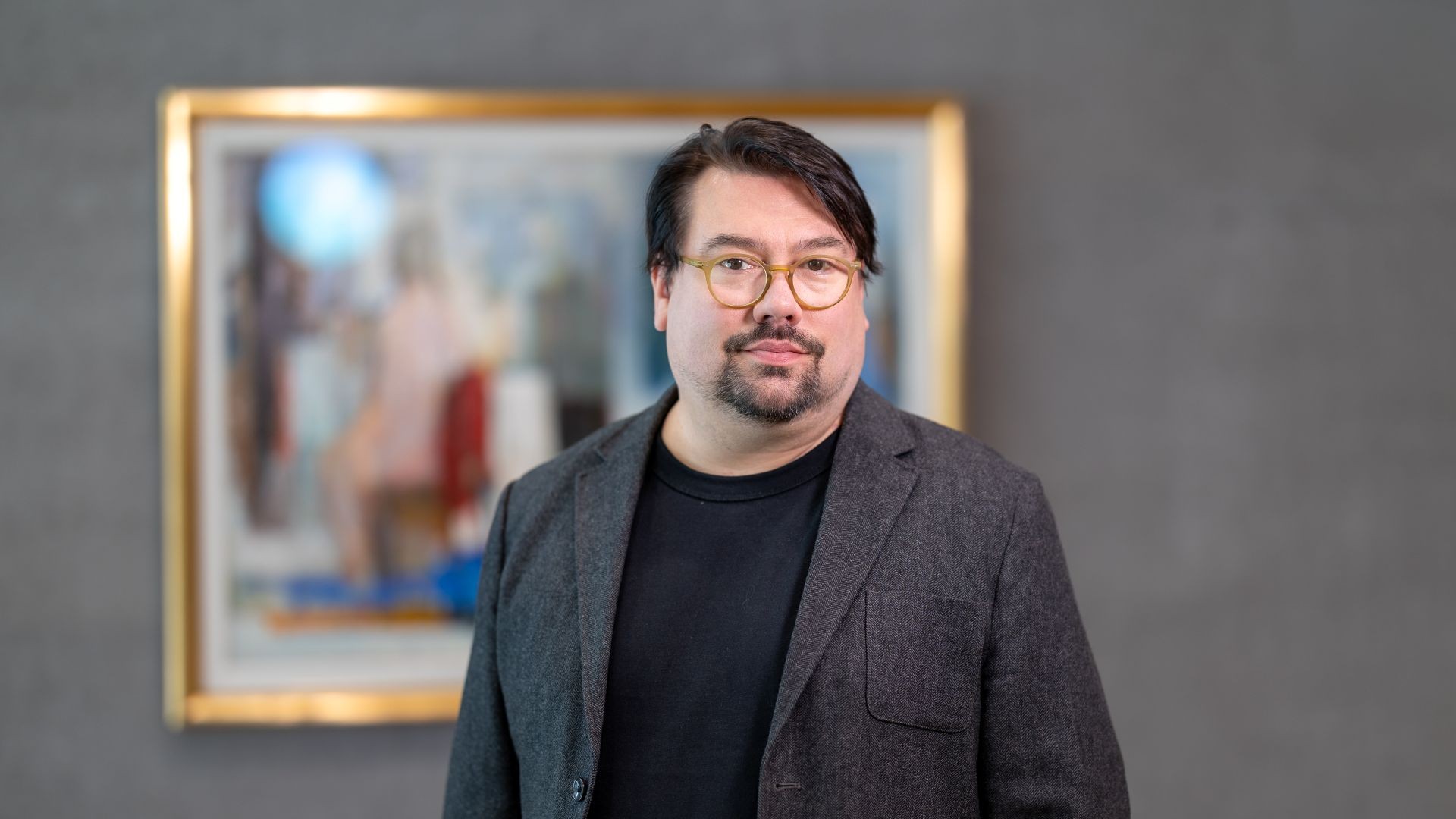 Veikko Halmetoja katsoo suoraan kameraan harmaassa puvuntakissa. Taustalla näkyy taulu.