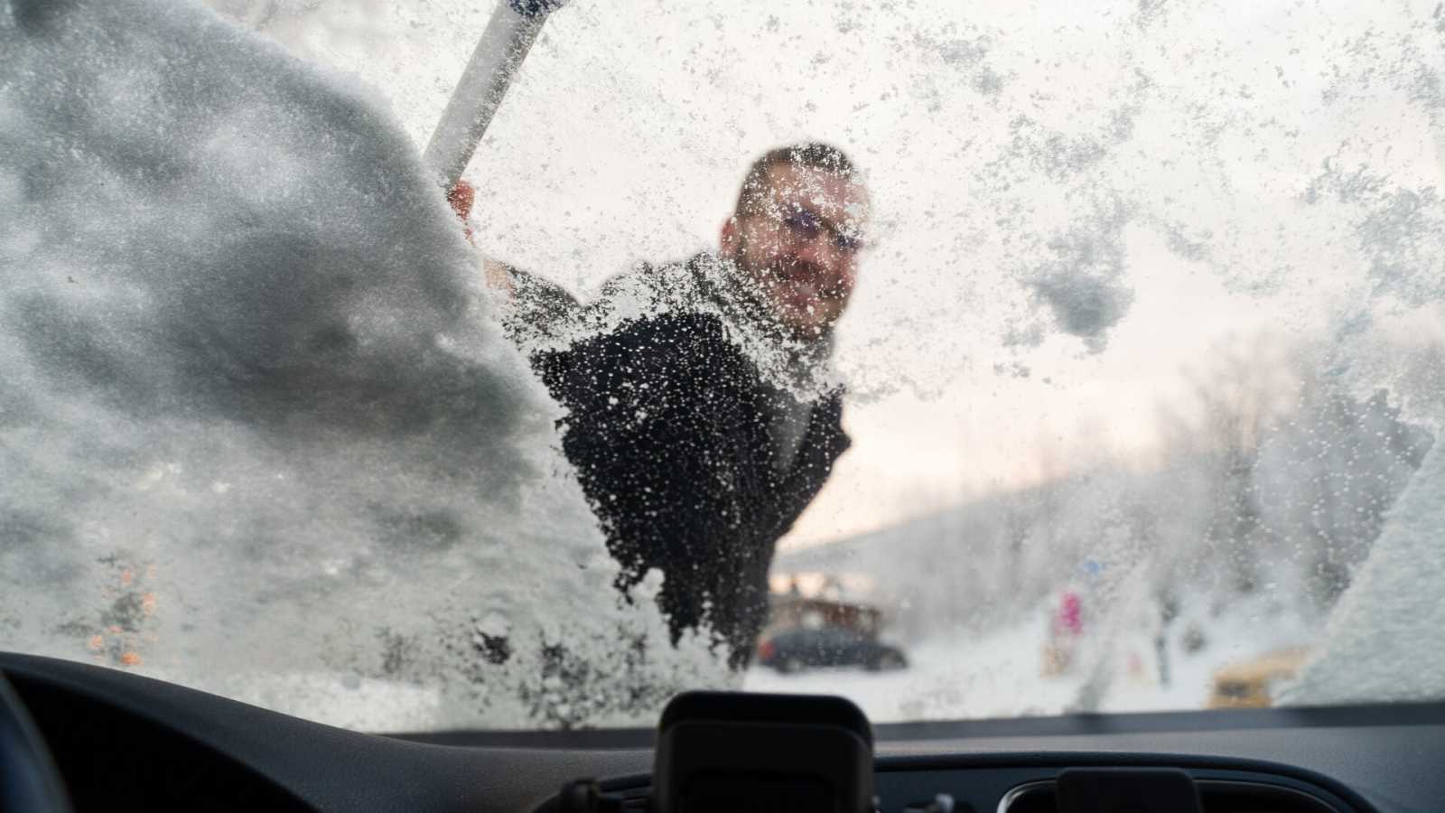 Hymyilevä mies pyyhkii lunta auton tuulilasista.