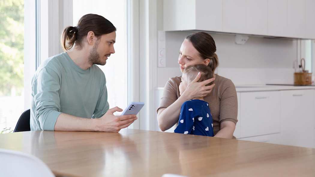 Perhe istuu pöydän ääressä. Äiti pitää sylissään kipeää lasta. Isä näppäilee puhelimella tietoja etälääkärille.