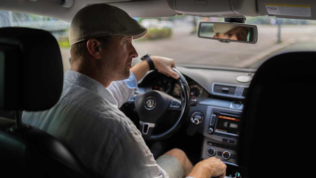 Mies ajaa autoa ja katsoo oikealle ikkunasta ulos siten, että hänen kasvonsa näkyvät peräpeilissä.