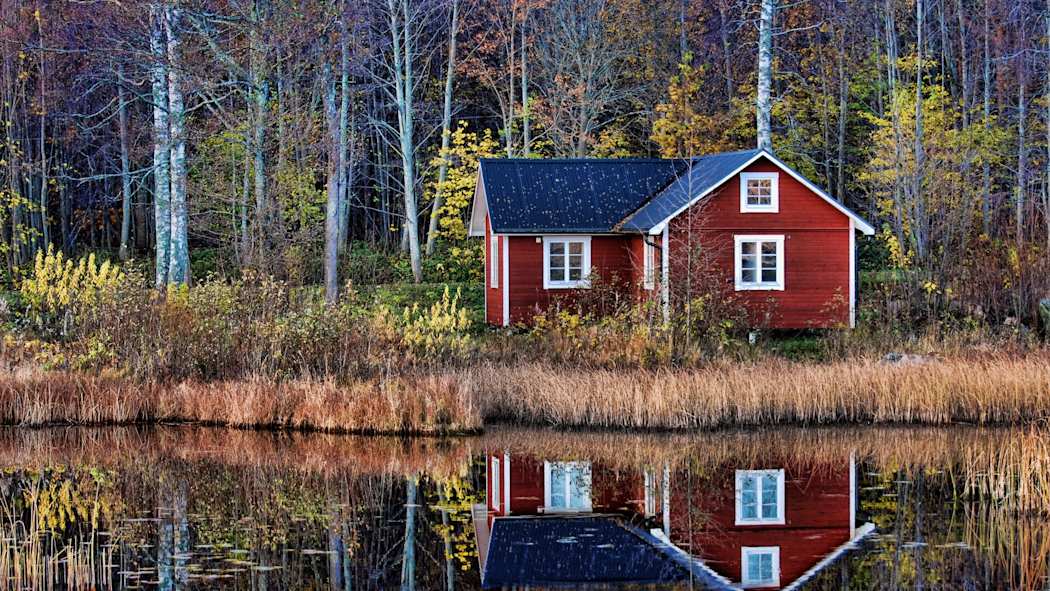 Punainen mökki metsän ja järven välissä. Puissa ja pensaissa näkyy ruskan värit.