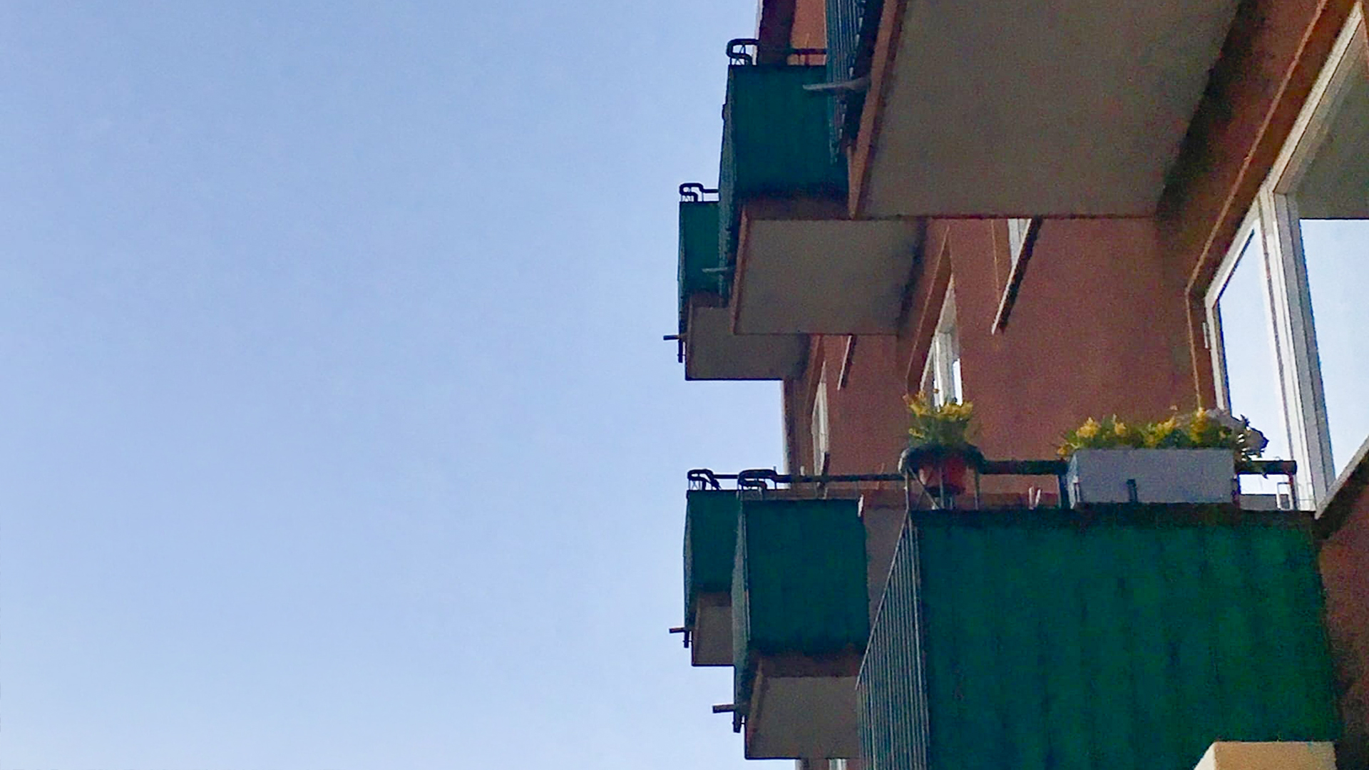Kerrostalon parvekkeet kuvattuna alhaalta päin. Alimmaisen parvekkeen laidalla on keltaisia kukkia.
