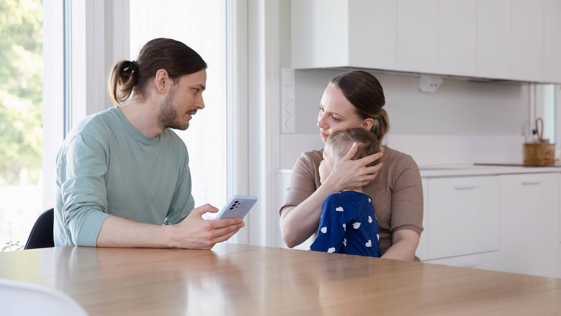 Nainen ja mies istuvat pöydän ääressä. Nainen halaa sairasta lasta sylissään, mies asioi vakuutusyhtiön terveyspalvelussa puhelimellaan.