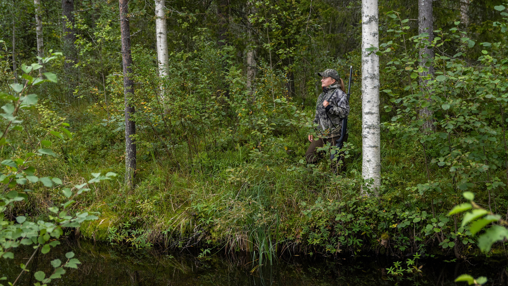 Metsästäjä seisoo joen varressa haulikko olallaan.