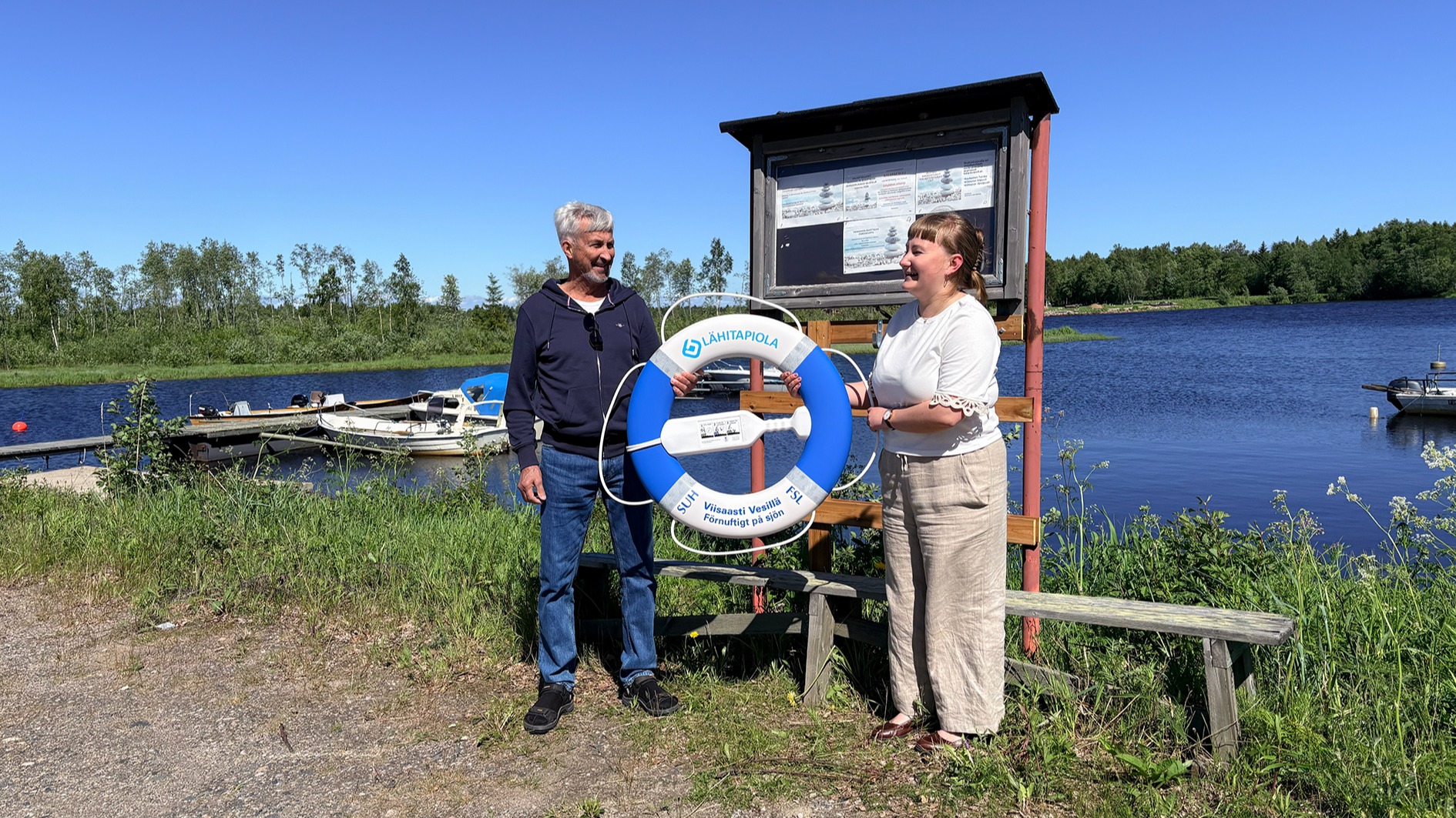 Mies ja nainen pitelevät LähiTapiolan sinivalkoista pelastusrengasta venelaitureiden edustalla. Aurinkoinen ja pilvetön taivas. 