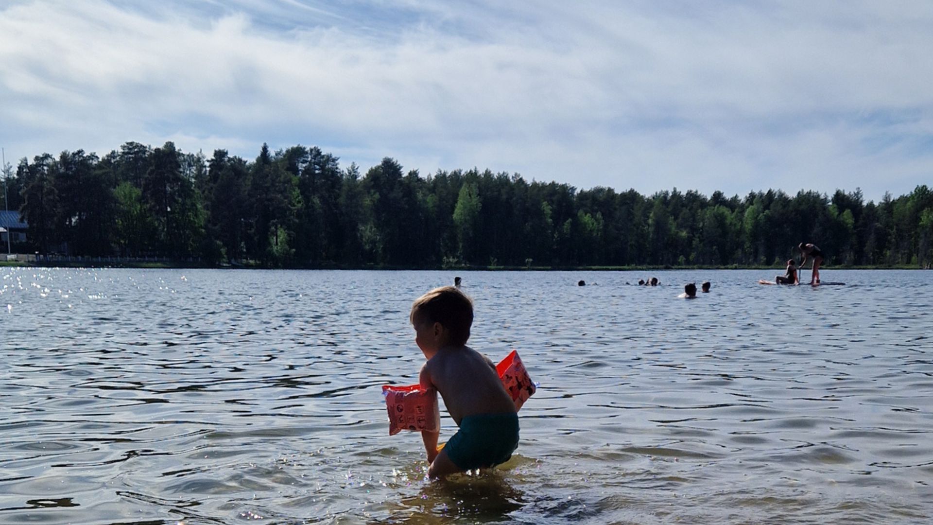 Kuvassa lapsi uimarannalla vedessä kesällä. Lapsella on uimakellukkeet. Taustalla näkyy metsämaisemaa. 