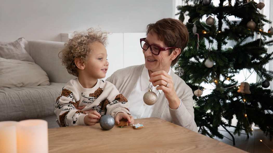 Isoäiti näyttää lapsenlapselleen joulukoristepalloa. Taustalla joulukuusi.