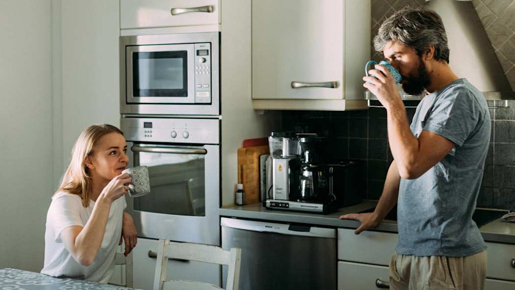 Paret dricker kaffe i köket. Kvinnan sitter vid bordet och mannen står vid köksbänken.