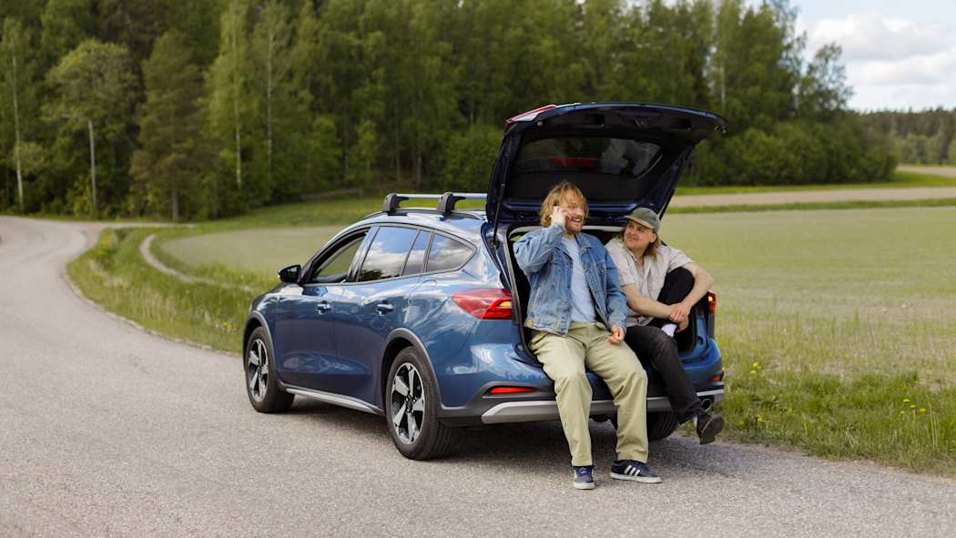 Kaksi miestä istuu auton takakontin reunalla. Auto on pysäytetty tien reunaan, toinen miehistä soittaa puhelimella.
