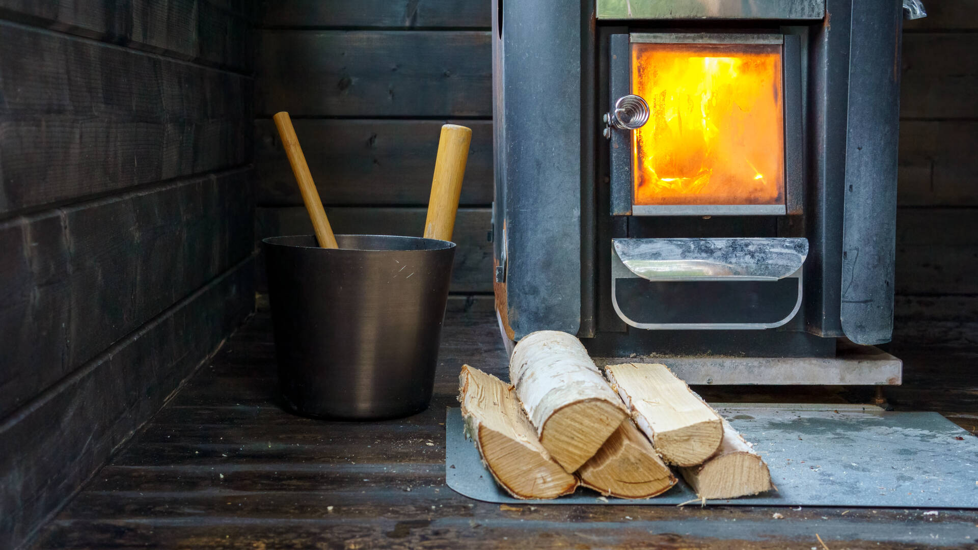 Saunan tulipesässä palaa tuli. Vieressä löylykiulu ja koivuhalkoja.
