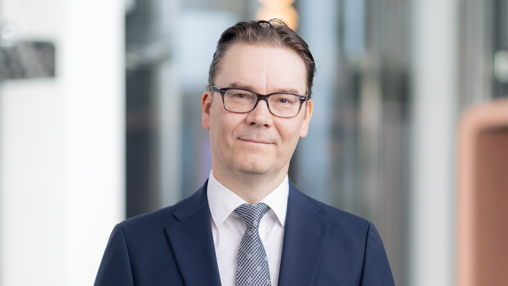 Potrettikuva tummansiniseen pukuun, valkoiseen paitaan ja kuviolliseen solmioon pukeutuneesta silmälaseja käyttävästä henkilöstä (Hannu Nummiaro). Tausta on pehmeästi rajattu toimistoympäristö.