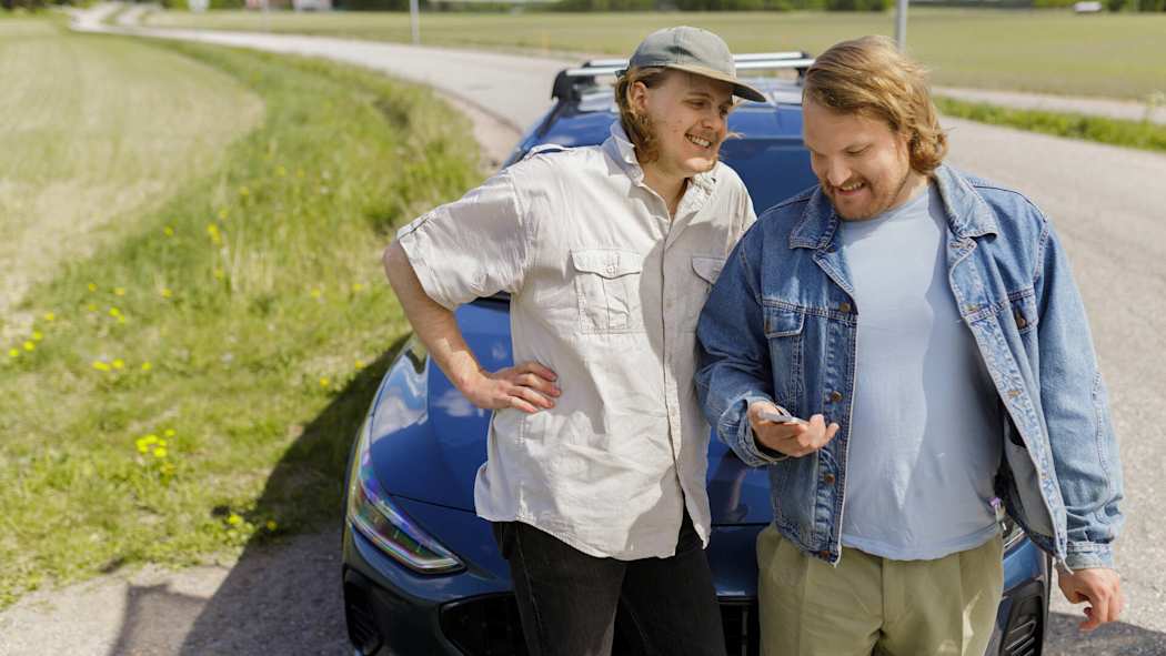Kaksi miestä on pysähtynyt tienvarteen. He seisovat autonsa edessä ja katsovat puhelinta.