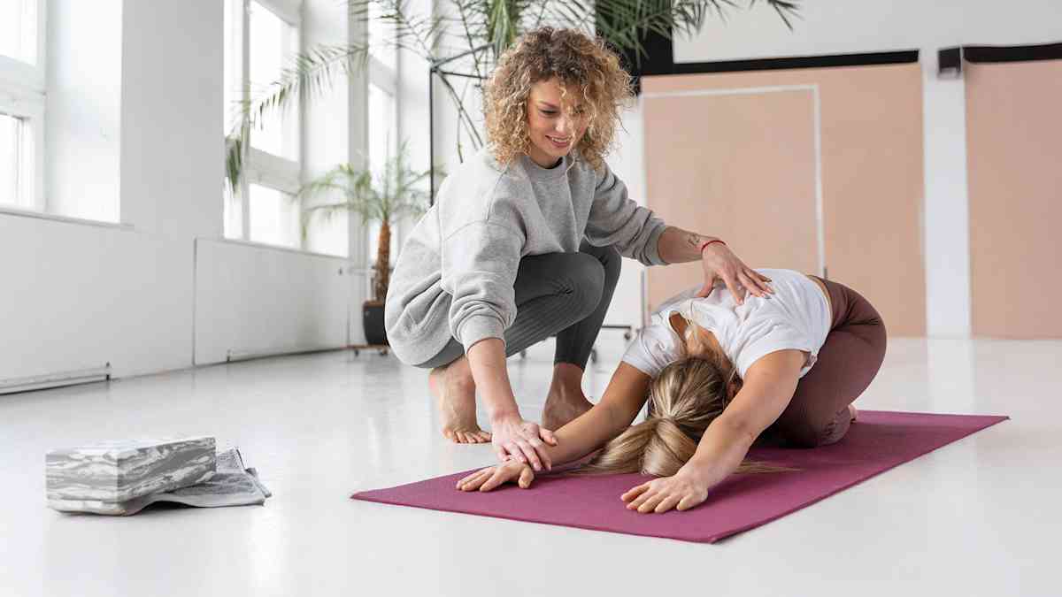 En lockhårig instruktör hjälper leende en kvinna som gör en yogastretch på en matta.