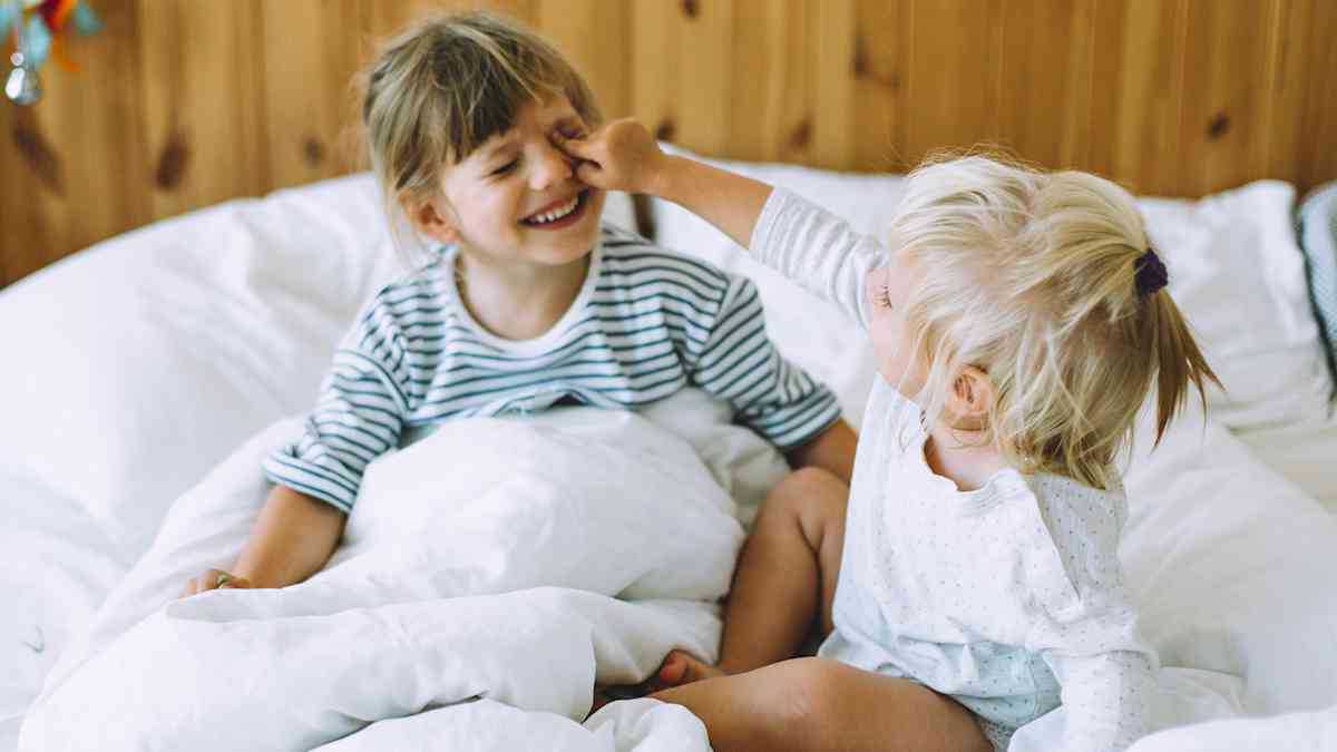 Kaksi pientä lasta leikkii ja nauraa valkoisissa lakanoissa sängyllä. Toinen koskettaa leikkisästi toisen kasvoja.