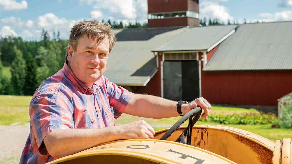 En rutklädd man sitter i en gul traktor på en bondgård. I bakgrunden syns en röd hall.