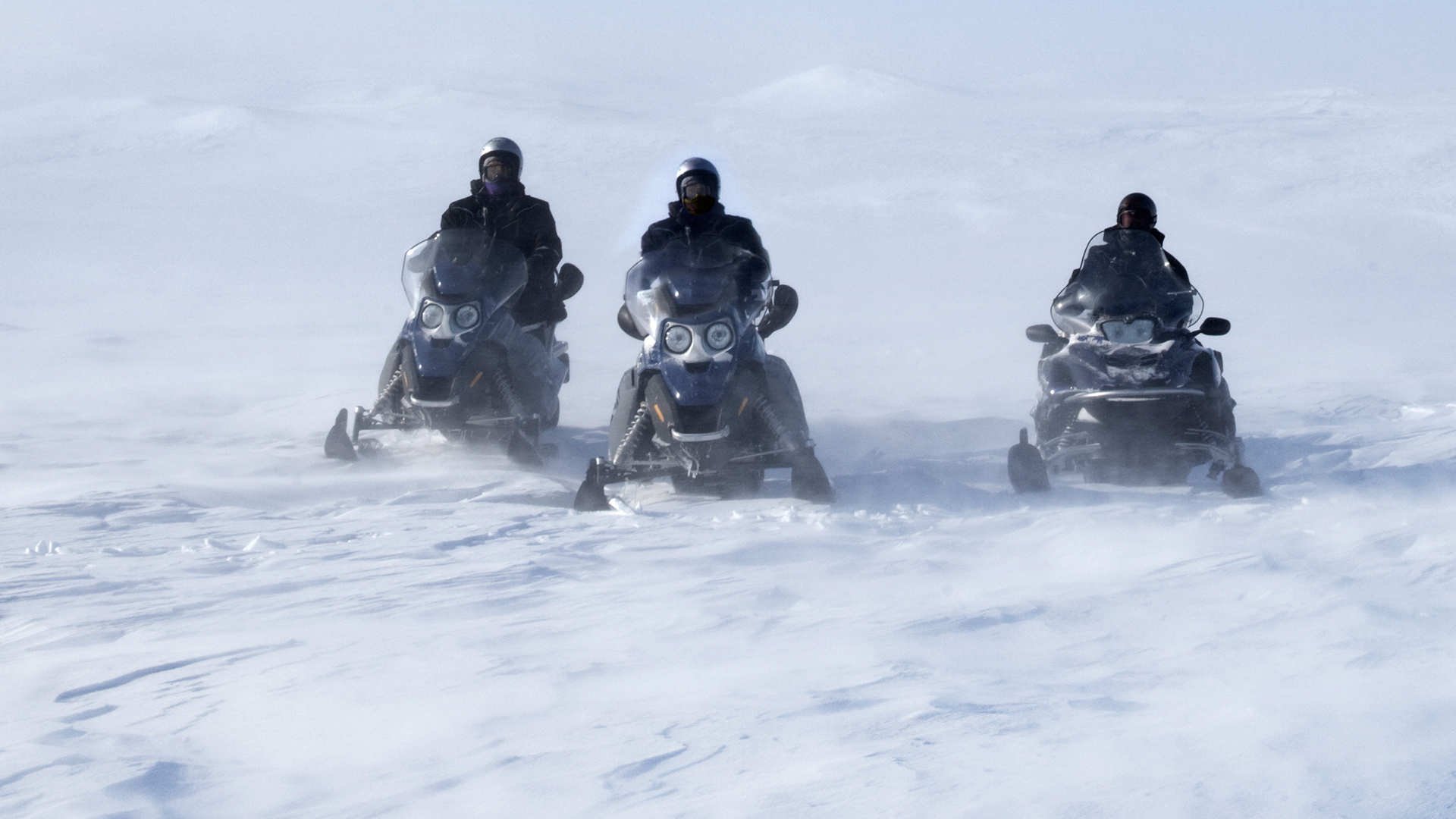 Tre personer kör snöskotrar sida vid sida i ett snöigt och vitt fjällandskap med hjälmarna på.